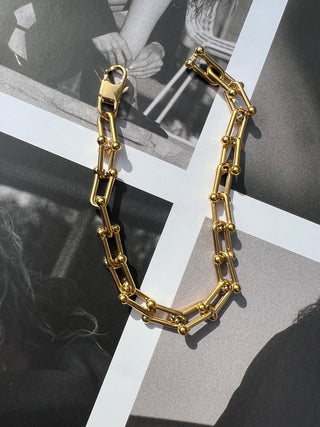 Gold rectangular-link chain bracelet on black-and-white photo background. Featured Product: Audrey Bracelet, SKU: BR-21289-001-RG