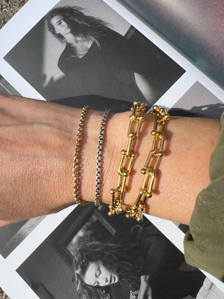 Wrist with layered gold and dark chain bracelets over an open magazine. Featured Product: Audrey Bracelet, SKU: BR-21289-001-RG